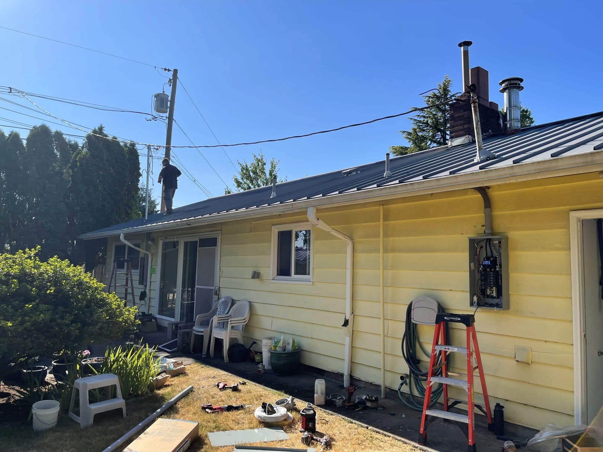 A sunny-day job on a yellow single-story house: a ladder set at the electrical panel on the side wall and a worker up on the metal roof.