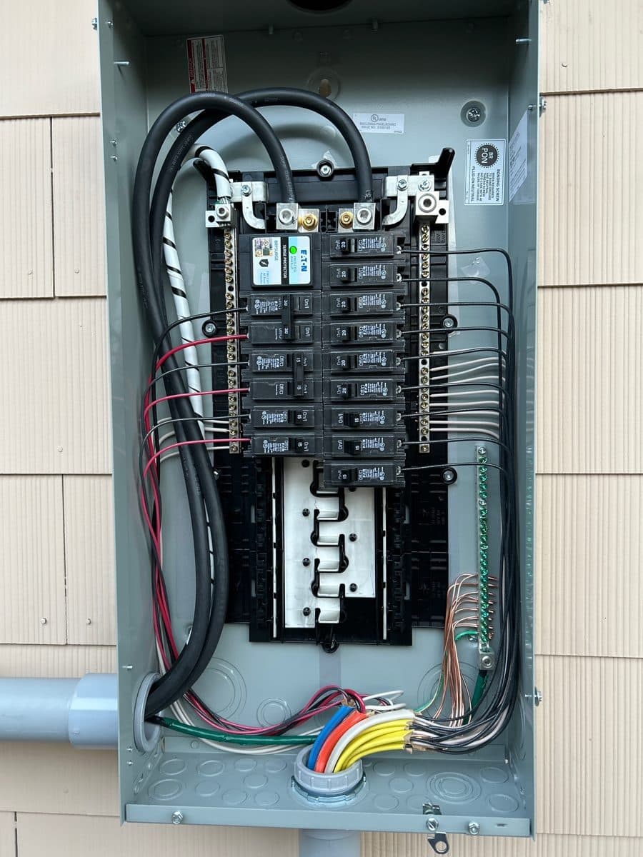A new panel in a gray outdoor enclosure on a shingle-sided house, with the new wires entering through the bottom.